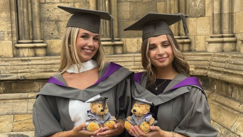 Two students wearing graduation gowns and hats, holding a small teddy bear dressed in a matching miniature graduation cap and gown.
